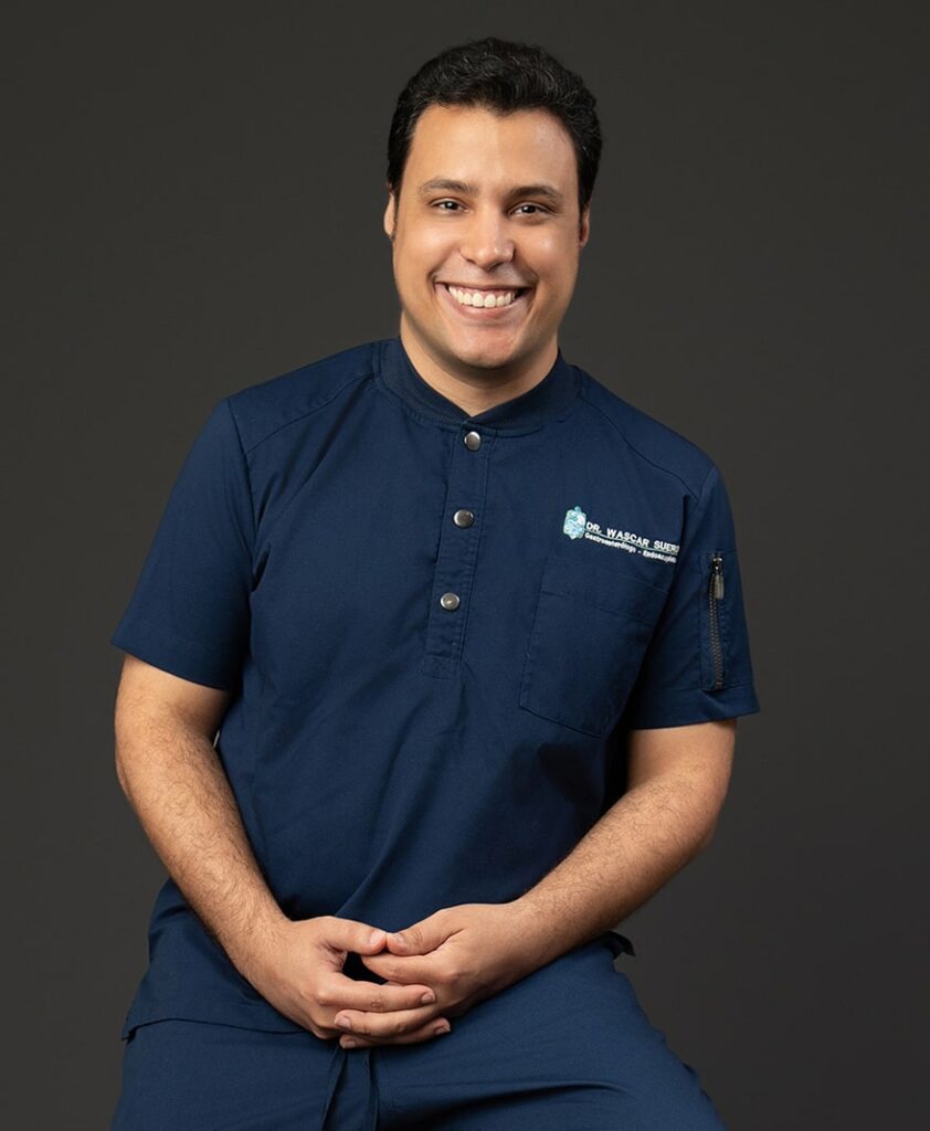 Foto de Wascar Suero, un doctor dominicano joven, sentado en una silla alta con las manos en las piernas, sonriendo a la cámara.