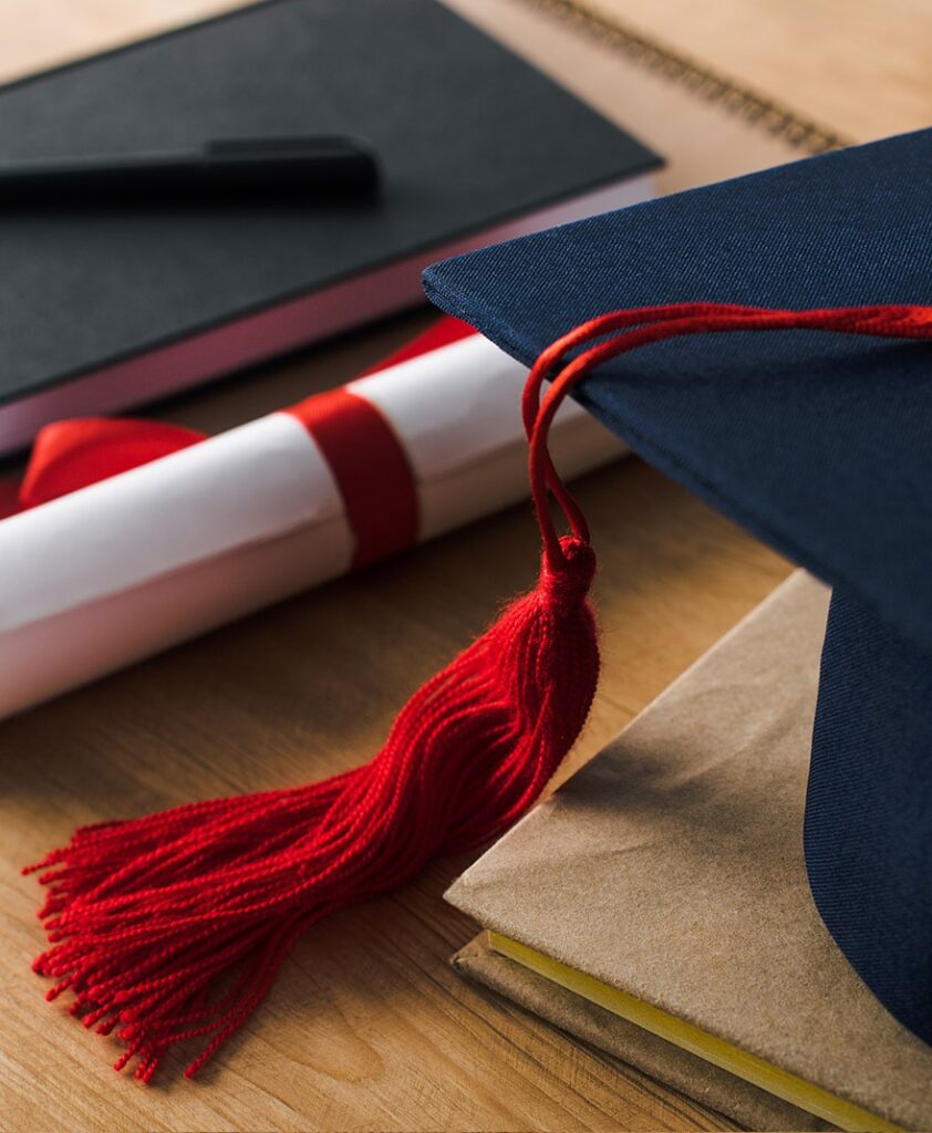 Diploma enrollado y atado con una cinta roja, junto a un birrete negro con una borla roja y varios cuadernos apilados sobre una mesa.